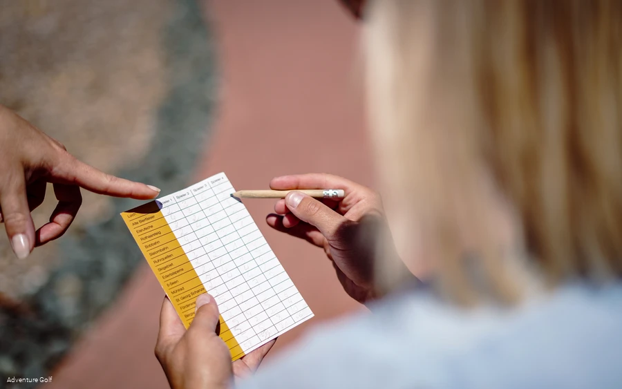 Eine Person schreibt mit einem Bleistift etwas auf den Zettel, wo die Schläge der Mitspieler erfasst werden.