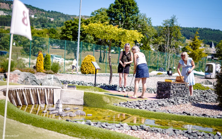 Drei Frauen spielen Golf auf einer Bahn mit der Edertalsperre und dem versunkenen Dorf im See.